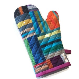 May include: A quilted oven mitt with a vibrant patchwork design. The mitt features a mix of colorful fabrics in shades of blue, orange, green, and purple, arranged in diagonal and diamond patterns. A magenta trim and a hanging loop complete the design.