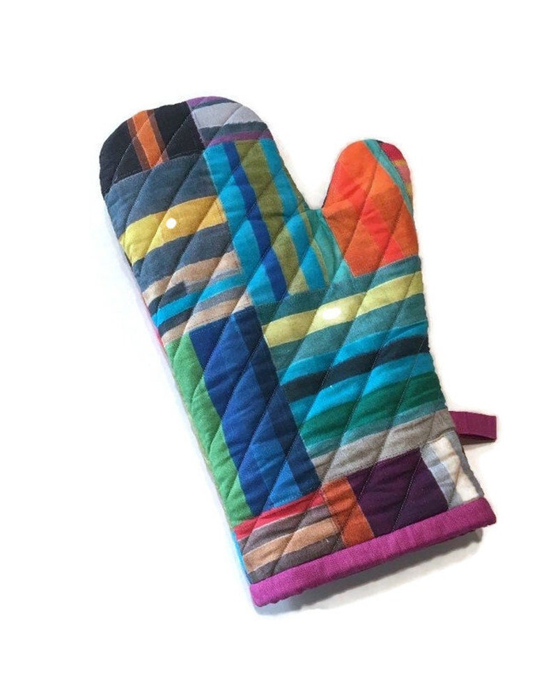 May include: A quilted oven mitt with a vibrant patchwork design. The mitt features a mix of colorful fabrics in shades of blue, orange, green, and purple, arranged in diagonal and diamond patterns. A magenta trim and a hanging loop complete the design.