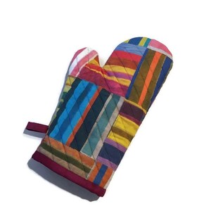 May include: A quilted oven mitt with a patchwork design. The mitt features a variety of colorful vertical stripes in shades of blue, red, yellow, and green. The edges are trimmed with a dark red fabric, and a small loop is attached for hanging.