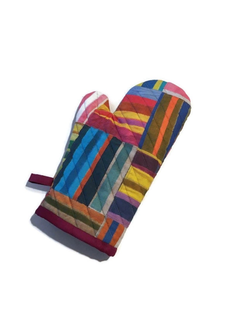 May include: A quilted oven mitt with a patchwork design. The mitt features a variety of colorful vertical stripes in shades of blue, red, yellow, and green. The edges are trimmed with a dark red fabric, and a small loop is attached for hanging.