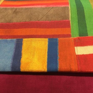May include: A colorful patchwork textile with a variety of rectangular and striped patterns. The design incorporates shades of red, orange, yellow, blue, green, brown, and purple. The bottom portion is a solid burgundy color.
