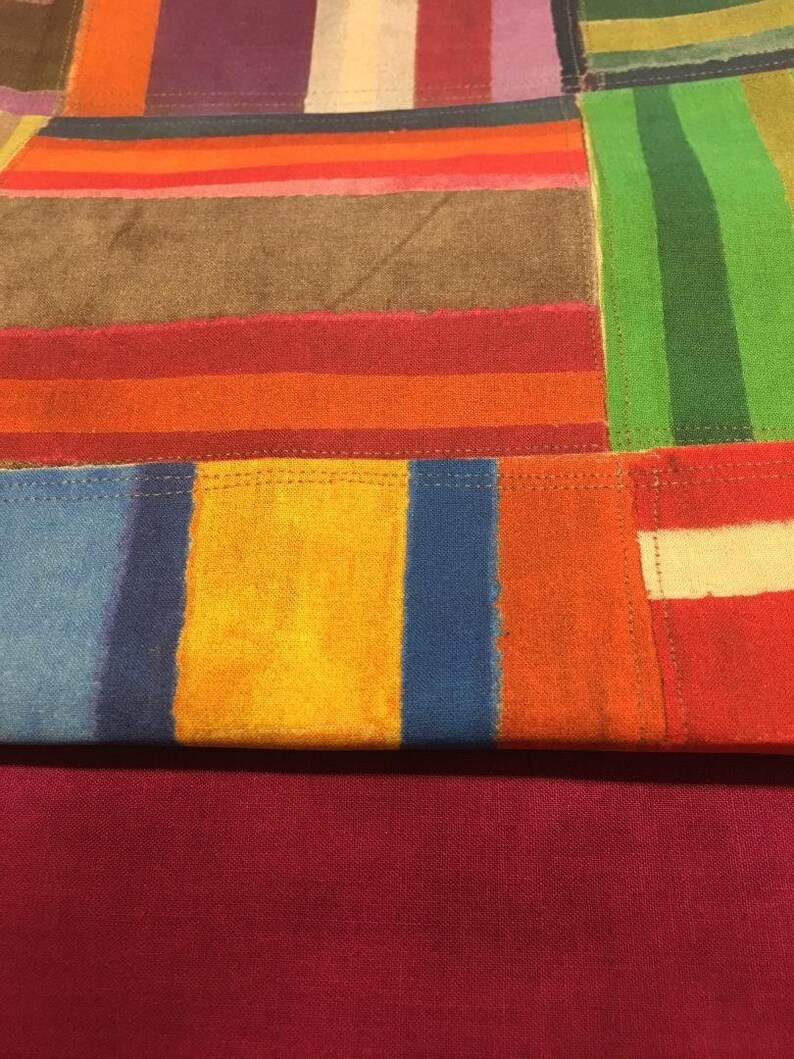May include: A colorful patchwork textile with a variety of rectangular and striped patterns. The design incorporates shades of red, orange, yellow, blue, green, brown, and purple. The bottom portion is a solid burgundy color.
