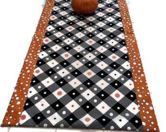 May include: A black and white checkered table runner with orange polka dot borders and a small orange pumpkin. The runner has a diamond pattern with white and orange accents. The table runner is a festive fall decoration.