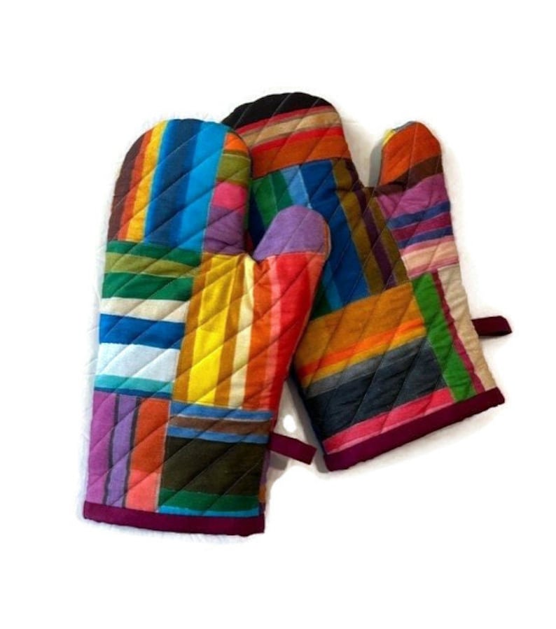 May include: A pair of quilted oven mitts with a patchwork design. The mitts feature a vibrant array of colors including blue, yellow, orange, and purple. The edges are trimmed with a dark pink binding.