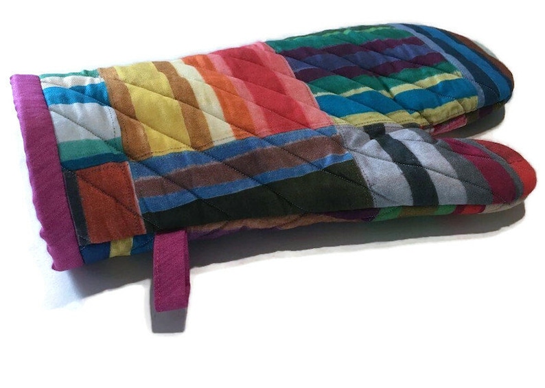 May include: A quilted oven mitt with a patchwork design. The mitt features a variety of colorful fabrics in stripes and squares, including red, orange, yellow, blue, and green. A magenta trim and hanging loop complete the design.