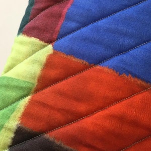 May include: Close-up of a quilted fabric with a vibrant, abstract design. The fabric features a patchwork of colours including blue, orange, green, and purple, with diagonal stitching. The design is reminiscent of a modern art piece.