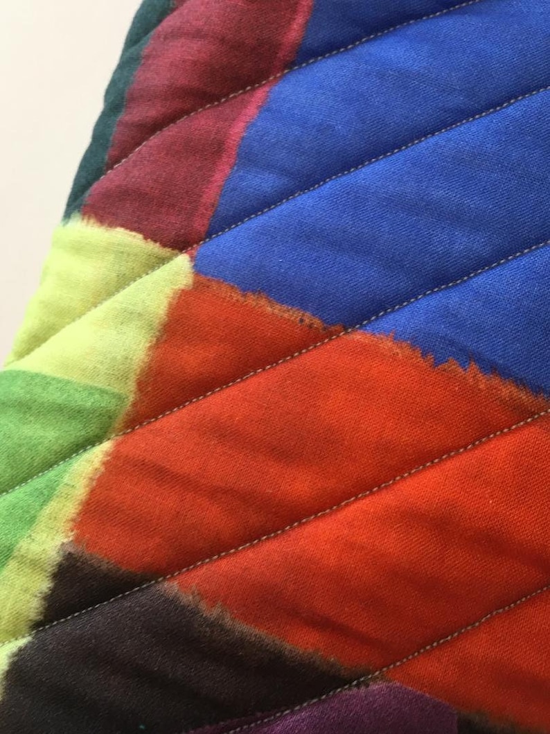 May include: Close-up of a quilted fabric with a vibrant, abstract design. The fabric features a patchwork of colours including blue, orange, green, and purple, with diagonal stitching. The design is reminiscent of a modern art piece.