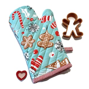 Christmas oven mitt features a light blue background with gingerbread cookies, mittens, and candy canes in shades of red, white, brown and black. A red and white stripe is used for lining and hanging loop. Cookie cutter props.