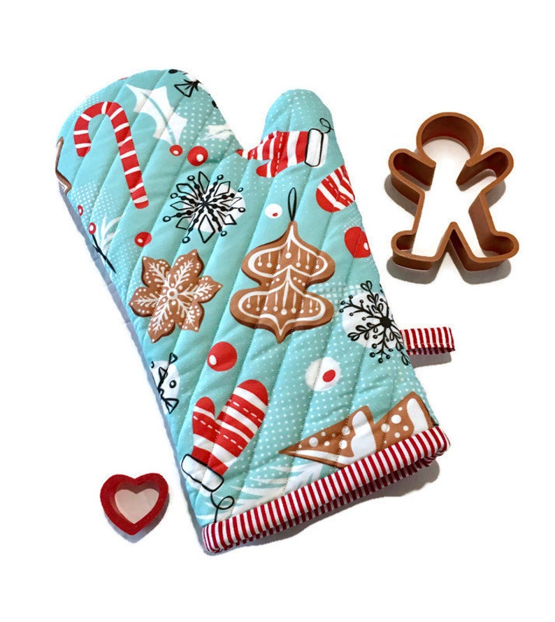 Christmas oven mitt features a light blue background with gingerbread cookies, mittens, and candy canes in shades of red, white, brown and black. A red and white stripe is used for lining and hanging loop. Cookie cutter props.