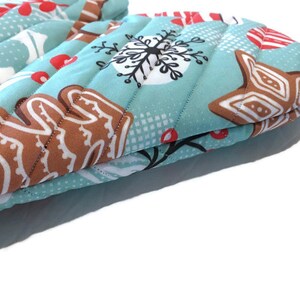 May include: A pair of quilted oven mitts with a light blue background and a repeating pattern of gingerbread men, snowflakes, and red berries. The mitts have a vertical quilted design.