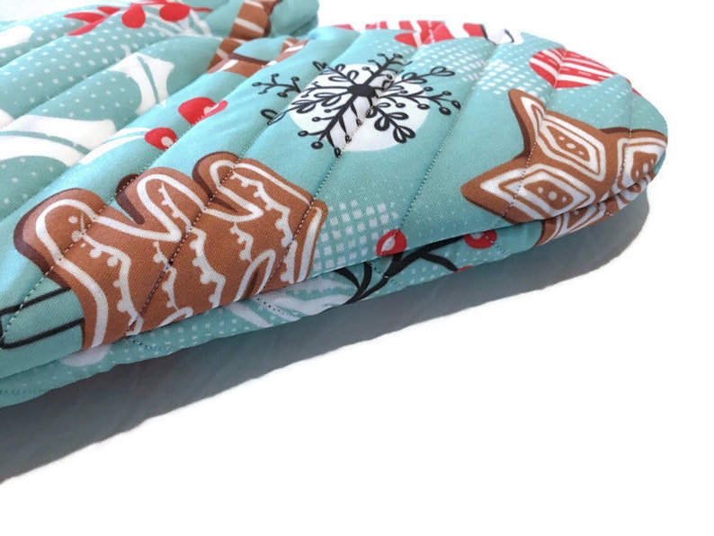 May include: A pair of quilted oven mitts with a light blue background and a repeating pattern of gingerbread men, snowflakes, and red berries. The mitts have a vertical quilted design.