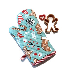 May include: A quilted oven mitt with a festive Christmas pattern in light blue, featuring gingerbread men, snowflakes, and candy canes. Includes a red heart and gingerbread man cookie cutters. The mitt has a red and white striped trim.