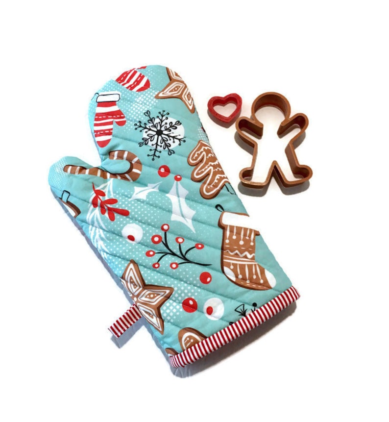 May include: A quilted oven mitt with a festive Christmas pattern in light blue, featuring gingerbread men, snowflakes, and candy canes. Includes a red heart and gingerbread man cookie cutters. The mitt has a red and white striped trim.