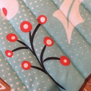 May include: Close-up of a quilted fabric with a light teal background and white polka dots. The fabric features a pattern of red berries on black stems and white floral shapes. Diagonal stitching adds texture to the design, suggesting a handmade item.