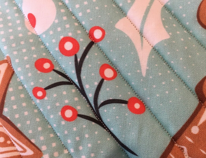 May include: Close-up of a quilted fabric with a light teal background and white polka dots. The fabric features a pattern of red berries on black stems and white floral shapes. Diagonal stitching adds texture to the design, suggesting a handmade item.