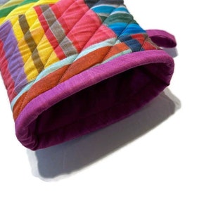 May include: A colorful quilted oven mitt with a vibrant striped pattern in shades of yellow, orange, pink, blue, and purple. The mitt has a quilted design and a magenta trim around the opening. A small loop is attached for hanging.