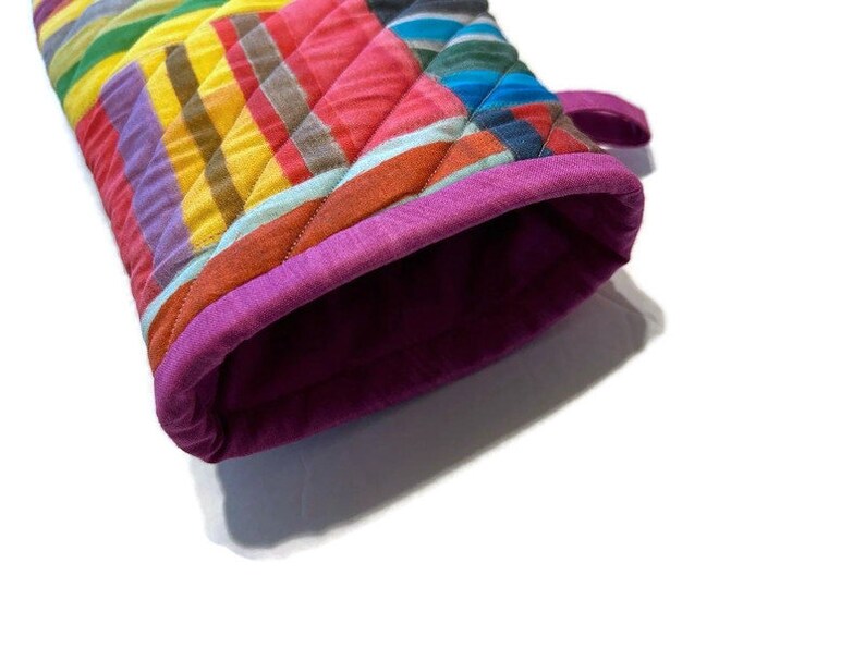 May include: A colorful quilted oven mitt with a vibrant striped pattern in shades of yellow, orange, pink, blue, and purple. The mitt has a quilted design and a magenta trim around the opening. A small loop is attached for hanging.