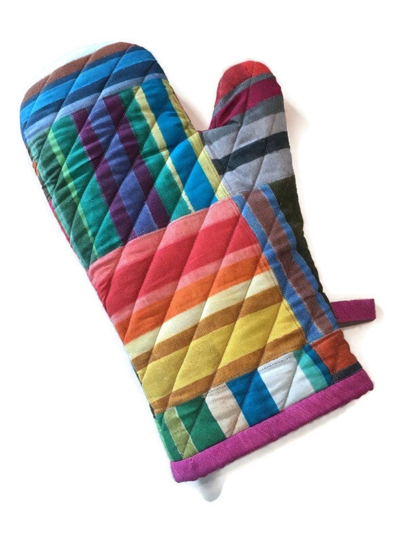 May include: A quilted oven mitt with a patchwork design. The mitt features a variety of colors, including blue, green, red, orange, and yellow, arranged in a striped and checkered pattern. The edges are trimmed with a bright pink binding.