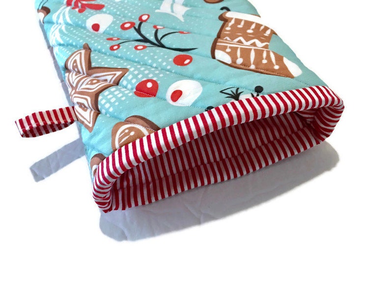 May include: A quilted oven mitt with a light blue background and a Christmas-themed pattern. The pattern includes gingerbread cookies, red berries, and white snowflakes. The cuff is red and white striped, and there is a red and white striped loop.