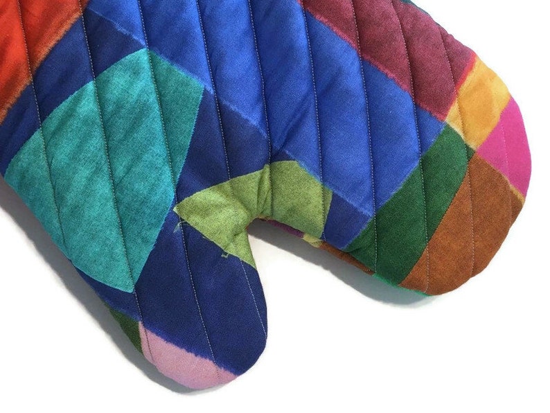 May include: A colourful quilted oven mitt with a patchwork design. The mitt features a variety of colours, including red, blue, green, and pink, arranged in a geometric pattern. The mitt is designed to protect hands from heat.