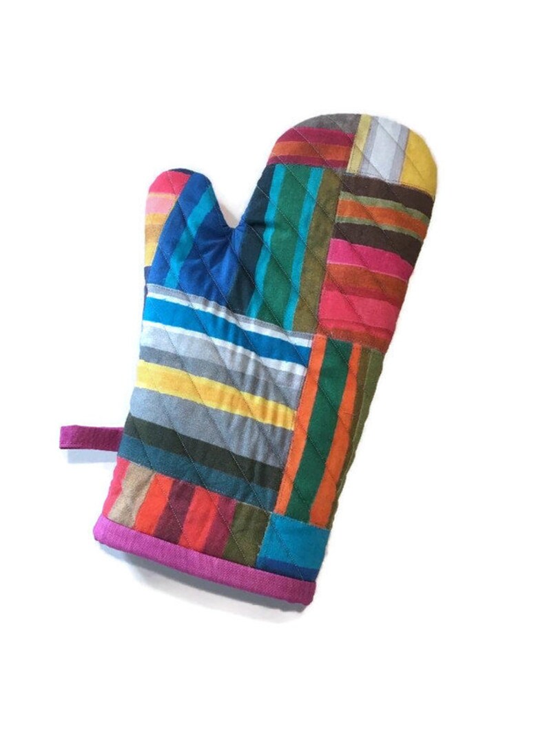 May include: A colorful oven mitt with a patchwork design. The mitt features a variety of vertical stripes in shades of blue, yellow, red, orange, and green. The cuff is trimmed with a bright pink fabric, and a pink loop is attached.