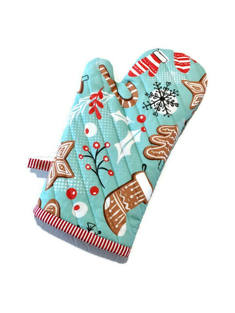 May include: A quilted oven mitt with a light blue background and a Christmas-themed pattern. The pattern includes gingerbread men, candy canes, snowflakes, and holly. The mitt has a red and white striped trim.