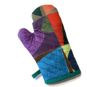 May include: A colourful patchwork oven mitt. The mitt is made up of various fabric pieces in shades of purple, blue, green, orange, and brown. It has a green hanging loop and green trim. Designed to protect hands from heat when handling hot cookware.