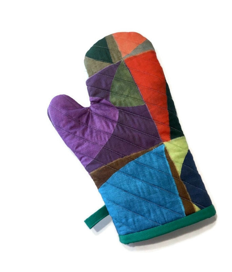 May include: A colourful patchwork oven mitt. The mitt is made up of various fabric pieces in shades of purple, blue, green, orange, and brown. It has a green hanging loop and green trim. Designed to protect hands from heat when handling hot cookware.
