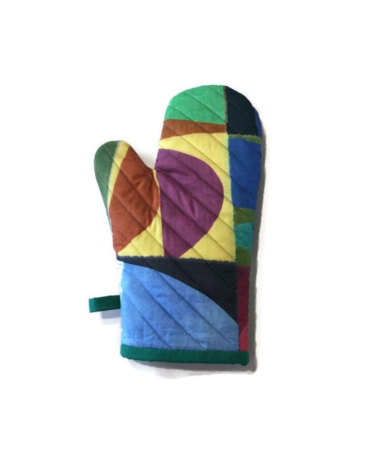 May include: A colourful quilted oven mitt with a patchwork design. The mitt features green, yellow, purple, blue, and brown geometric shapes. It has a green loop for hanging and a green trim around the edge.