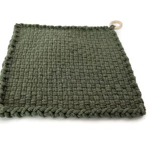 May include: A square, olive green woven potholder with a braided edge and a wooden ring for hanging. The potholder is made of a thick, textured material.