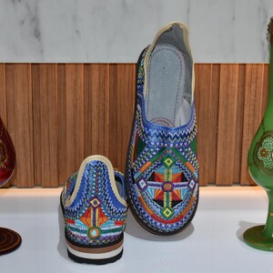 Puede incluir: Zapatos bordados con un vibrante dise&ntilde;o geom&eacute;trico en azul, naranja y verde. Flanqueando los zapatos hay dos jarrones decorativos de cristal, uno rojo y otro verde, con detalles dorados. Los zapatos miden unos 24 cm de largo.