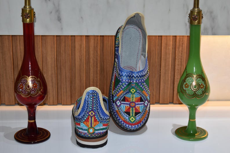 Puede incluir: Zapatos bordados con un vibrante dise&ntilde;o geom&eacute;trico en azul, naranja y verde. Flanqueando los zapatos hay dos jarrones decorativos de cristal, uno rojo y otro verde, con detalles dorados. Los zapatos miden unos 24 cm de largo.