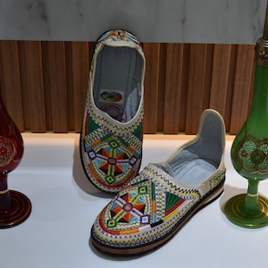 Puede incluir: Par de zapatos sin cordones bordados con patrones geom&eacute;tricos coloridos. Los zapatos son beige con detalles en verde, rojo, amarillo y azul. Dos botellas decorativas de vidrio con dise&ntilde;os ornamentados flanquean los zapatos.