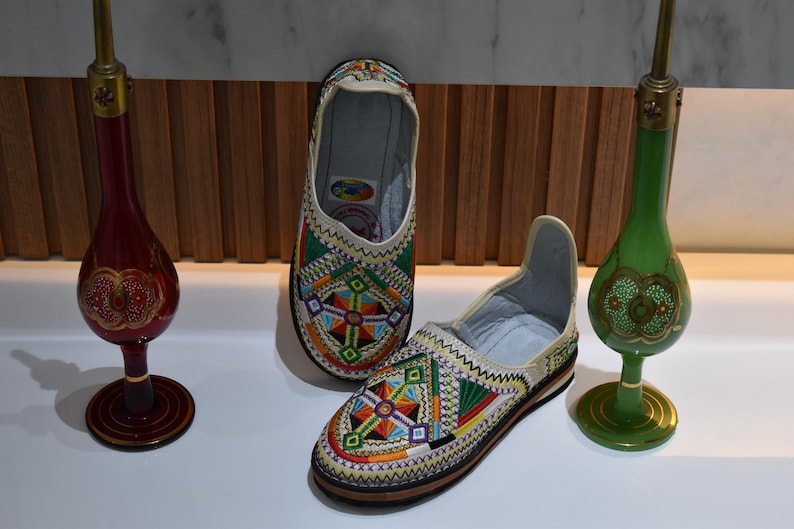 Puede incluir: Par de zapatos sin cordones bordados con patrones geom&eacute;tricos coloridos. Los zapatos son beige con detalles en verde, rojo, amarillo y azul. Dos botellas decorativas de vidrio con dise&ntilde;os ornamentados flanquean los zapatos.