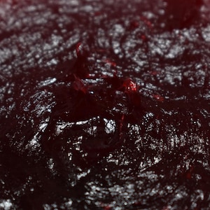May include: Close-up of a dark red, textured food item, possibly a jam or sauce. The surface has a glossy sheen with visible variations in color and texture. The image highlights the rich, deep color and the natural, uneven surface.