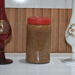 May include: A clear plastic jar with a red lid, filled with a granular substance, sits between two decorative glass vases. The vases are a deep red and white, with gold and floral designs. The background is a wooden panel.