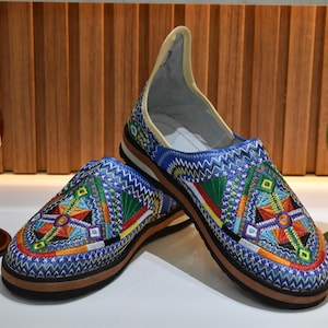 Puede incluir: Par de zapatos sin cordones bordados en azul con patrones geom&eacute;tricos coloridos. Los zapatos tienen suela marr&oacute;n e interior color crema. Jarrones decorativos de cristal rojo y verde flanquean los zapatos.