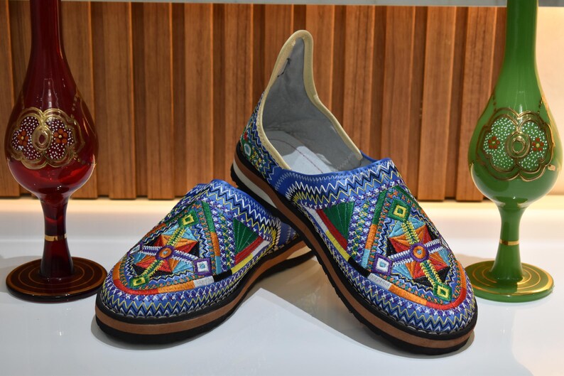Puede incluir: Par de zapatos sin cordones bordados en azul con patrones geom&eacute;tricos coloridos. Los zapatos tienen suela marr&oacute;n e interior color crema. Jarrones decorativos de cristal rojo y verde flanquean los zapatos.