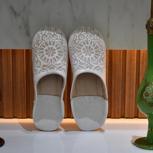 Puede incluir: Un par de zapatillas de cuero blanco con un diseño floral beige. Las zapatillas se exhiben entre una botella de vidrio decorativa roja y verde. El fondo presenta un panel de madera y una superficie blanca.