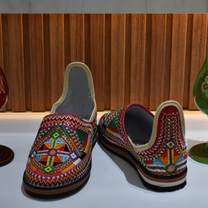 Puede incluir: Zapatillas bordadas con dise&ntilde;os geom&eacute;tricos coloridos en rojo, amarillo, verde y azul. Dos jarrones decorativos de cristal, uno rojo y otro verde, flanquean los zapatos. Las zapatillas miden aproximadamente 25 cm.