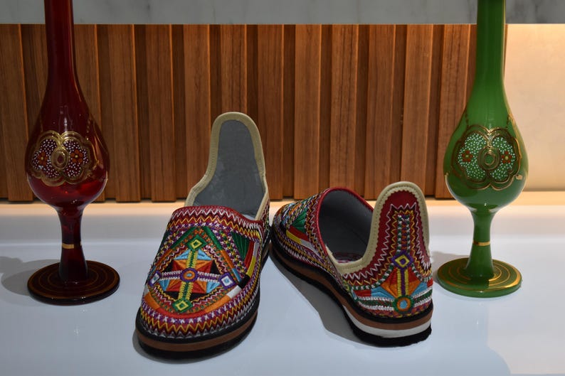 Puede incluir: Zapatillas bordadas con dise&ntilde;os geom&eacute;tricos coloridos en rojo, amarillo, verde y azul. Dos jarrones decorativos de cristal, uno rojo y otro verde, flanquean los zapatos. Las zapatillas miden aproximadamente 25 cm.