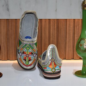 Puede incluir: Zapatos bordados con un dise&ntilde;o geom&eacute;trico y parte superior plateada. Flanqueando los zapatos hay dos botellas decorativas de vidrio, una roja y otra verde, con detalles dorados. El conjunto se exhibe sobre una superficie blanca.