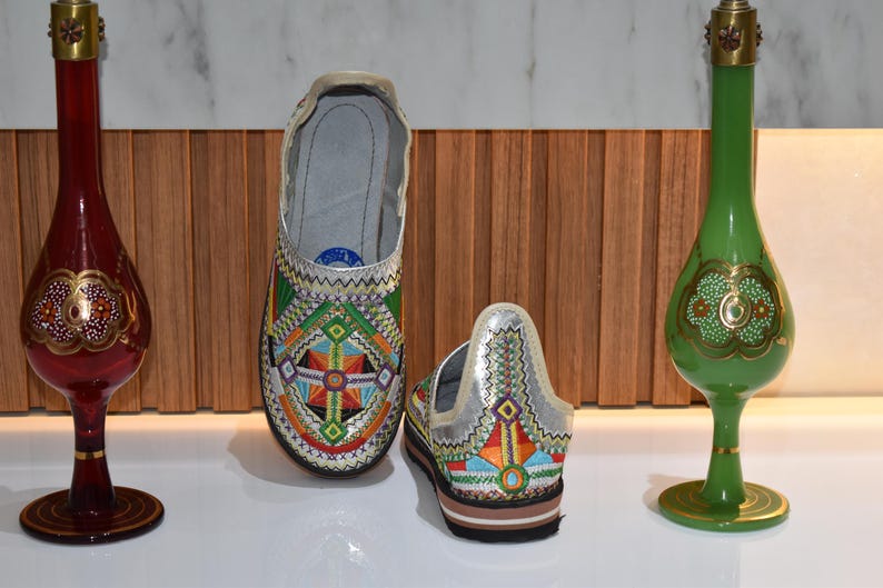 Puede incluir: Zapatos bordados con un dise&ntilde;o geom&eacute;trico y parte superior plateada. Flanqueando los zapatos hay dos botellas decorativas de vidrio, una roja y otra verde, con detalles dorados. El conjunto se exhibe sobre una superficie blanca.