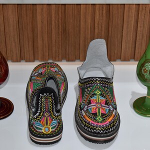 Puede incluir: Dos pares de coloridas zapatillas bordadas con base negra y patrones intrincados. Dos botellas decorativas de vidrio, una roja y otra verde, con detalles dorados, se encuentran a ambos lados de las zapatillas. El fondo es un panel de madera.