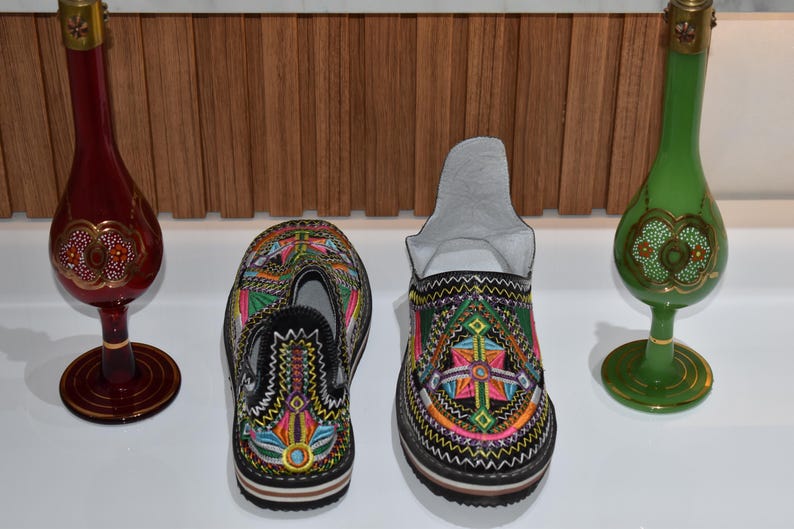 Puede incluir: Dos pares de coloridas zapatillas bordadas con base negra y patrones intrincados. Dos botellas decorativas de vidrio, una roja y otra verde, con detalles dorados, se encuentran a ambos lados de las zapatillas. El fondo es un panel de madera.