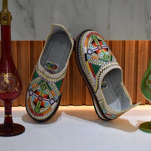 Puede incluir: Un par de zapatos sin cordones bordados con patrones geom&eacute;tricos coloridos en rojo, verde, azul y amarillo. Los zapatos se exhiben con dos jarrones decorativos de vidrio, uno rojo y otro verde, con detalles dorados.