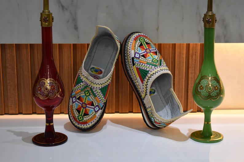 Puede incluir: Un par de zapatos sin cordones bordados con patrones geom&eacute;tricos coloridos en rojo, verde, azul y amarillo. Los zapatos se exhiben con dos jarrones decorativos de vidrio, uno rojo y otro verde, con detalles dorados.