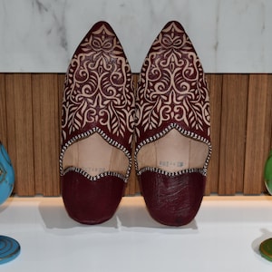 Puede incluir: Un par de babuchas de cuero burdeos con intrincados diseños florales blancos. Se exhiben entre dos jarrones decorativos de cristal, uno azul y otro verde, con detalles dorados. El fondo presenta un panel de madera y una superficie blanca.