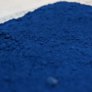 May include: Close-up of a pile of vibrant, deep blue powder. The powder has a fine, granular texture and is slightly uneven, with small clumps visible. The color is a rich, saturated blue, suggesting a pigment or dye.
