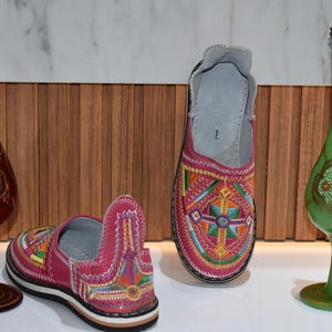 Puede incluir: Dos pares de zapatos rosas bordados sin cordones con patrones geom&eacute;tricos coloridos. Una jarra decorativa de vidrio rojo y verde con detalles dorados a cada lado de los zapatos.
